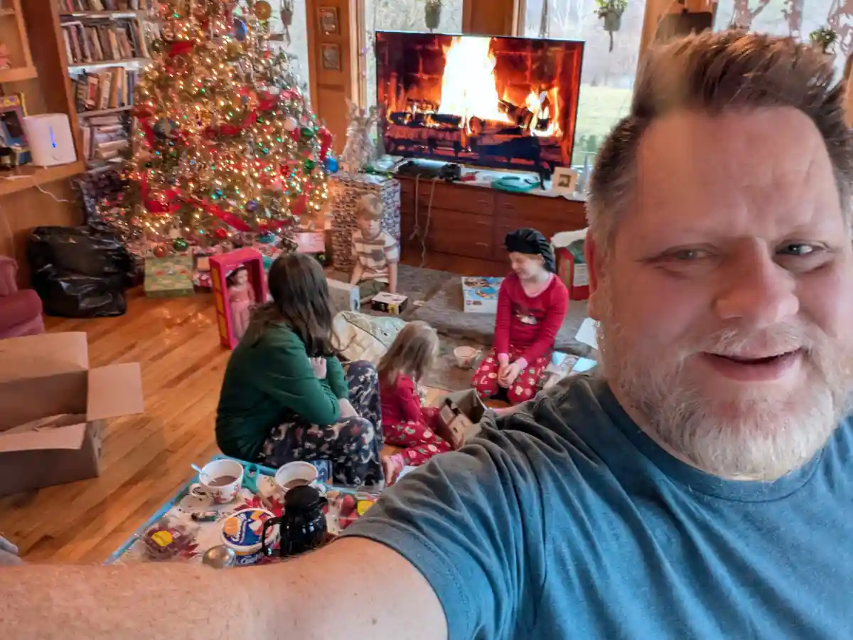 A man takes a selfie in a living room decorated for Christmas, with a lit tree, presents, and several children in pajamas…