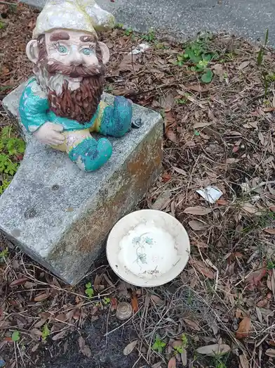 A weathered garden gnome with peeling paint sits on a concrete block outdoors, next to a dirty, empty white bowl with a…