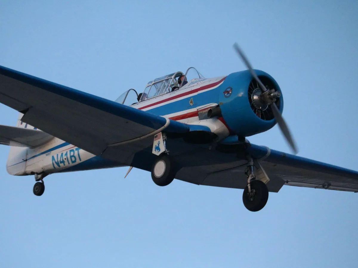 A blue, white, and red vintage single-engine propeller airplane with landing gear down flies against a clear blue sky. The…