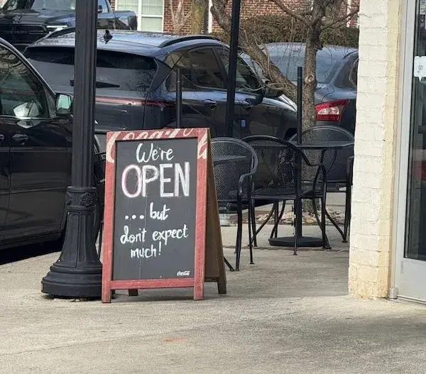 A red-framed chalkboard sign outside a business reads 'We're OPEN ... but don't expect much!' in white chalk. The sign…