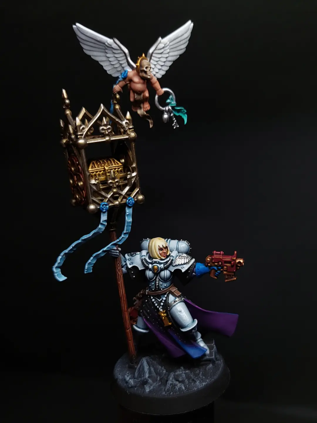 A painted miniature of a female space marine in silver armor holding a large ornate staff topped with a winged skull figure…