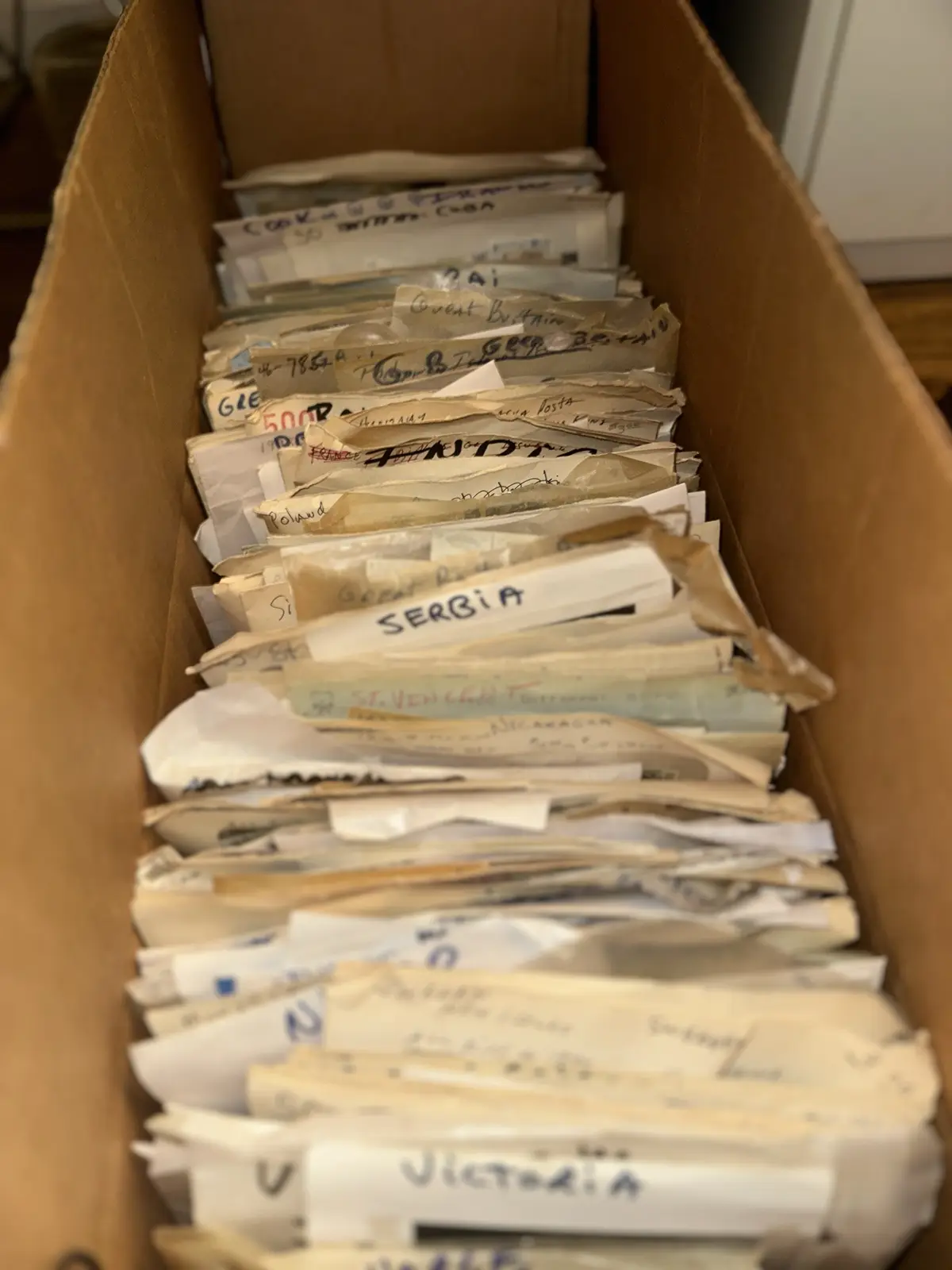 A long cardboard box is filled with numerous small, individually wrapped packages, each labeled with handwritten country…