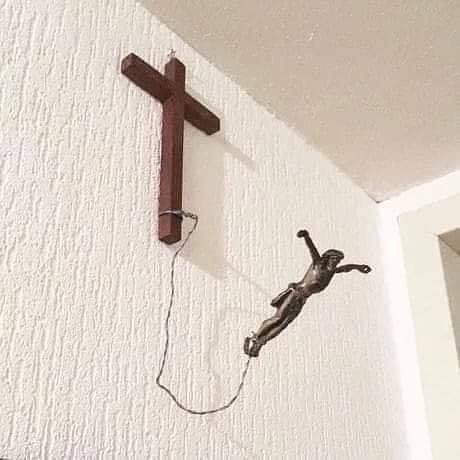 A wooden cross is mounted on a white textured wall, with a bronze statue of Jesus hanging from it by a thin rope, appearing…