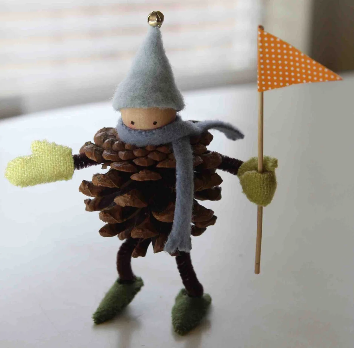 A handmade figurine crafted from a pinecone, wearing a grey felt hat with a bell, a grey scarf, and green felt mittens and…