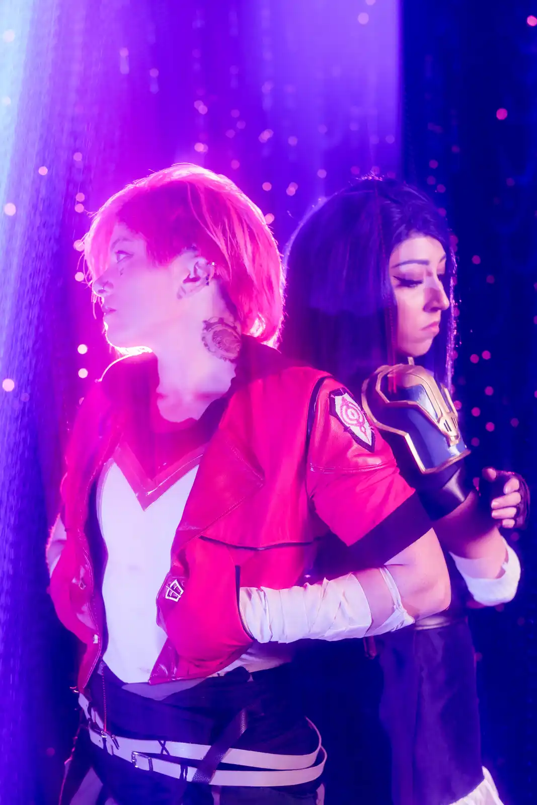 Two people in cosplay stand back-to-back under purple and pink stage lighting; the person on the left, with short pink hair…