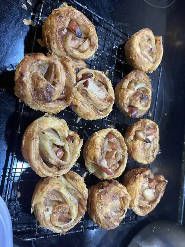 A close-up photo shows several golden-brown, flaky apple turnovers cooling on a black wire rack. The pastries have a spiral…