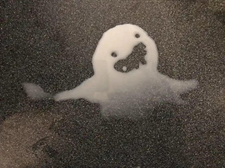 A white, ghost-shaped puddle of milk or similar liquid sits on a dark, speckled surface, with two small black dots for eyes…