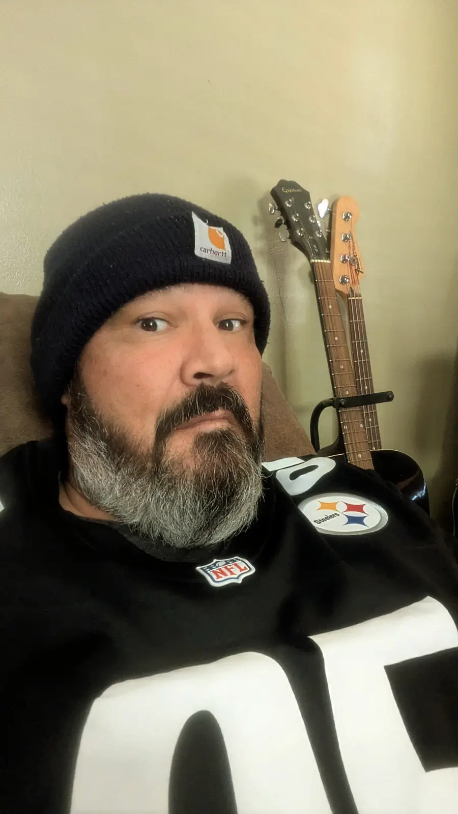 A man with a graying beard and mustache wears a black Pittsburgh Steelers NFL jersey and a black Carhartt beanie while…