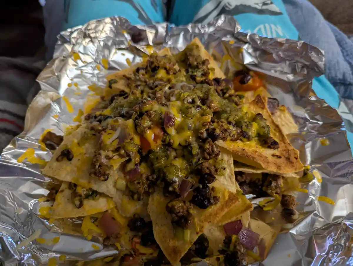 A close-up photo shows a generous serving of loaded nachos on crumpled aluminum foil, topped with melted cheese, ground…