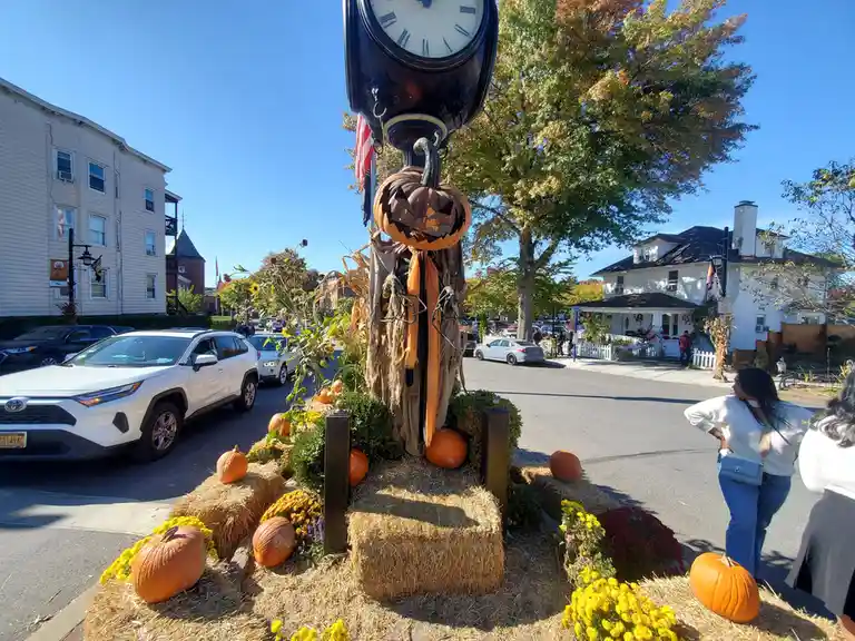 A street scene features a decorative clock with a carved jack-o'-lantern pumpkin as its face, surrounded by hay bales…