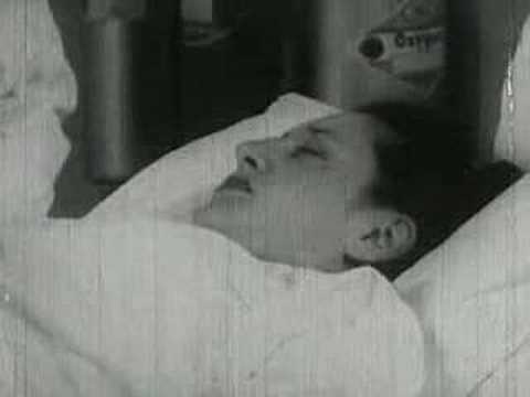 A grainy black-and-white photo shows a person lying in bed, eyes closed, appearing to be asleep or resting. The image has a…