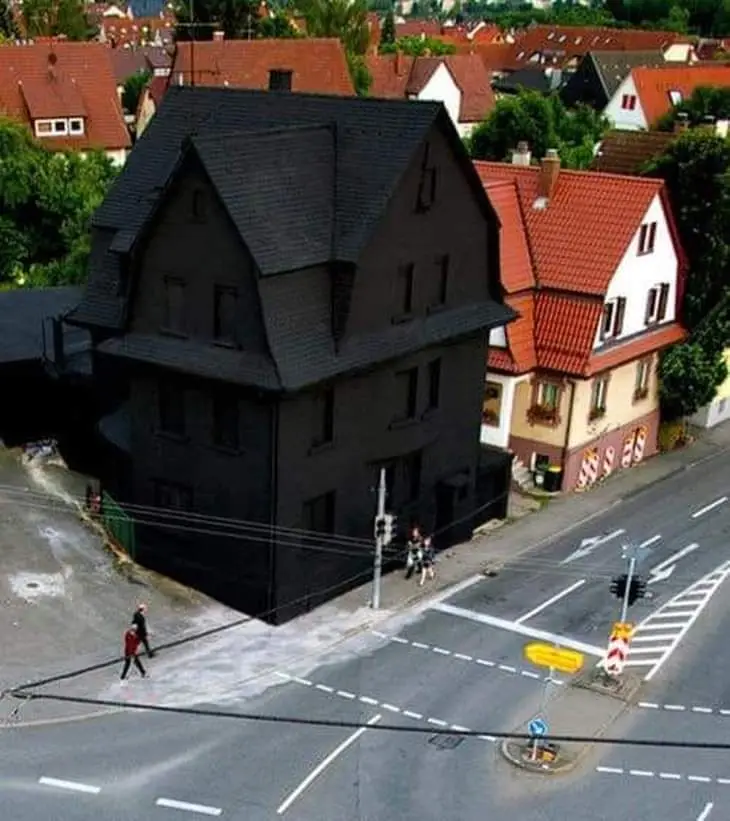 an aerial view shows a large, dark-colored house that appears to be physically consuming the street corner and sidewalk next…
