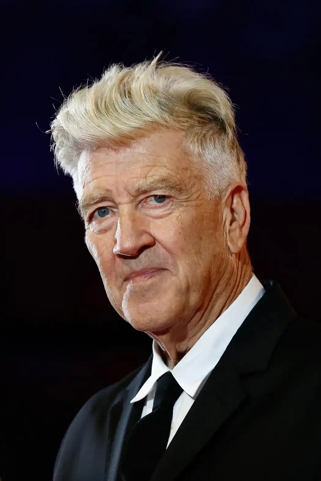 A close-up photo of David Lynch, an older man with distinctive spiky blonde hair, wearing a black suit and white shirt…