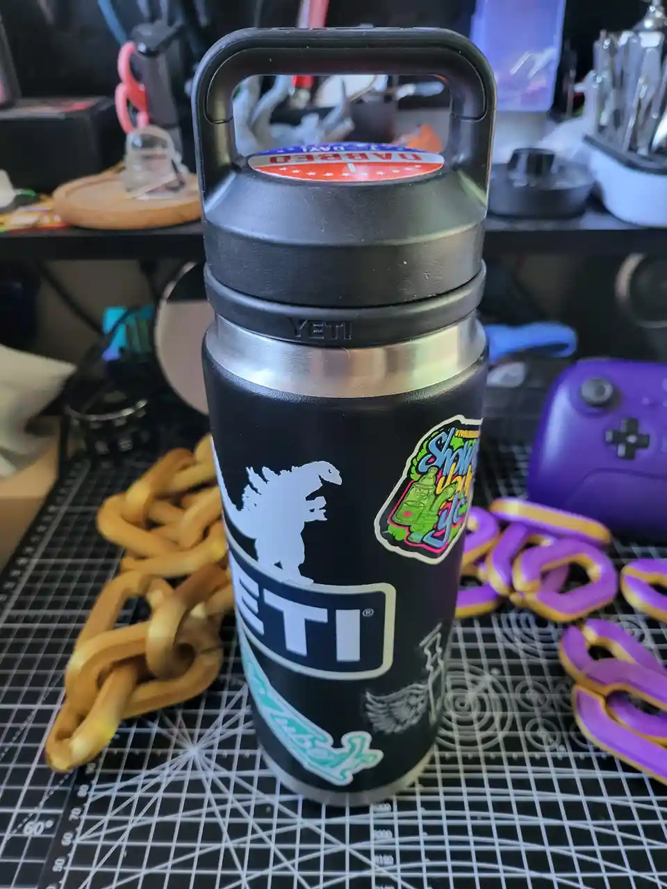 A black Yeti water bottle with multiple stickers, including a Godzilla silhouette and a 'Shawty' design, sits on a black…
