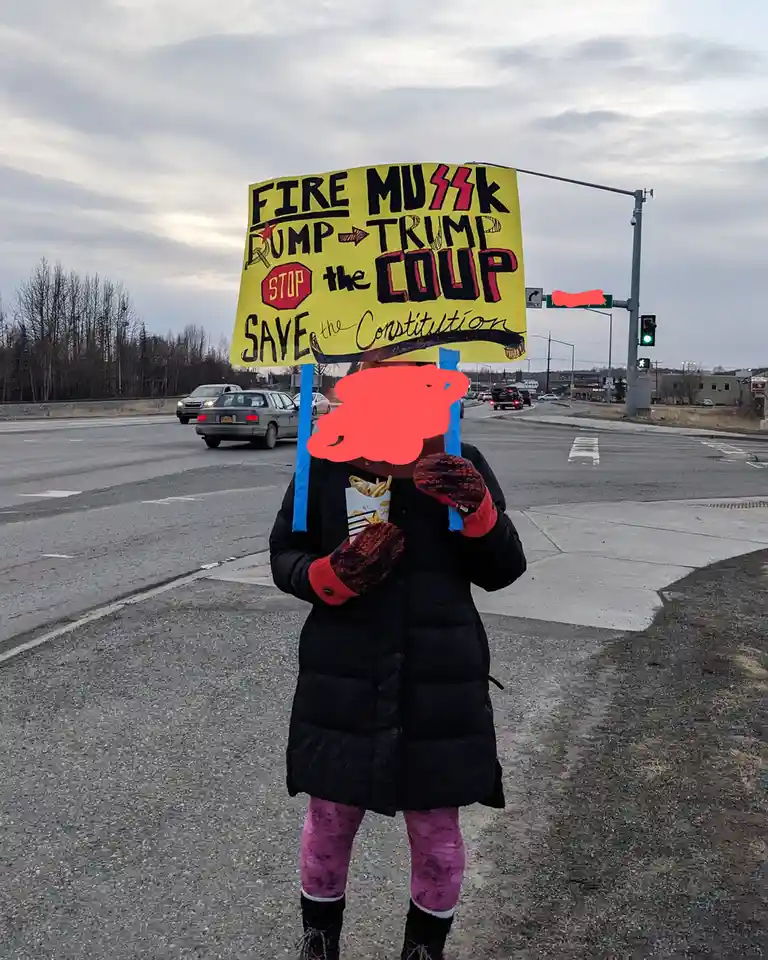 A person wearing a black puffer coat, pink leggings, and mittens stands at a street corner holding a large yellow protest…