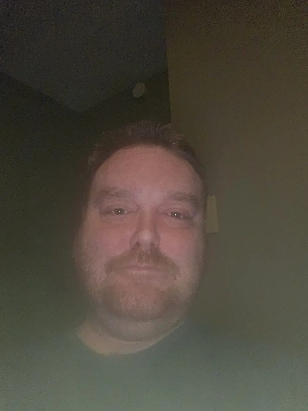 A low-resolution, dimly lit selfie of a man with a mustache and beard, looking directly at the camera with a neutral…