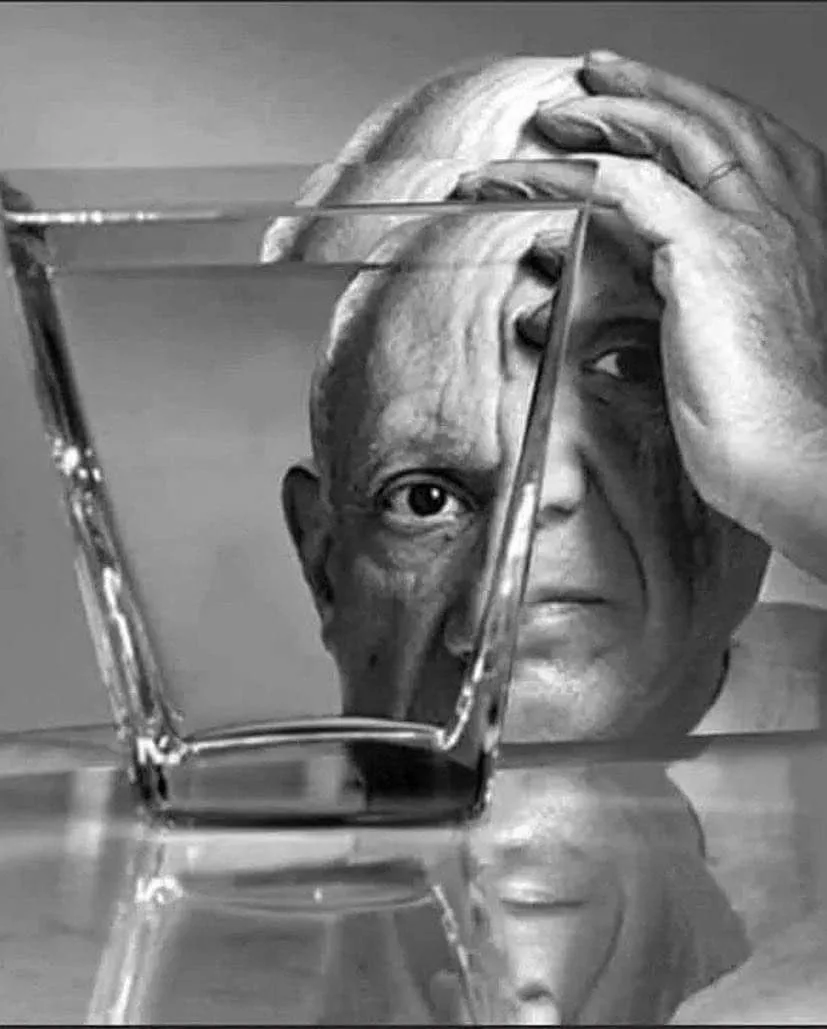 A black-and-white photo shows an older man with his hands on his head, partially obscured by a glass of water in front of…