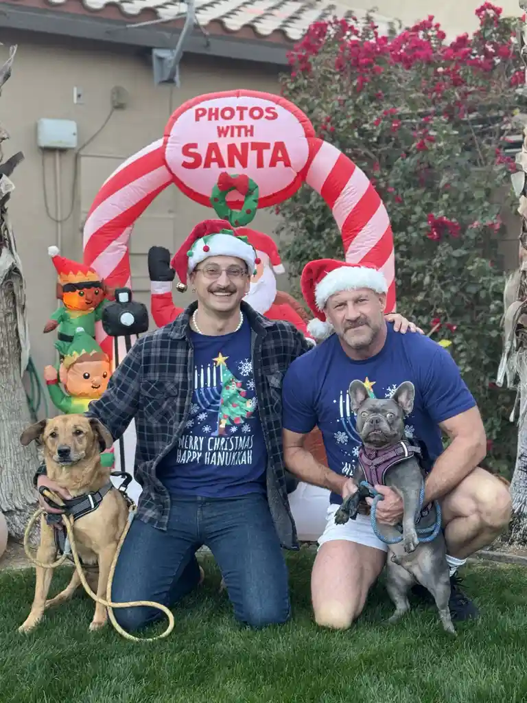 Two men in Santa hats kneel on grass in front of a large inflatable candy cane arch labeled 'PHOTOS WITH SANTA'. One man…