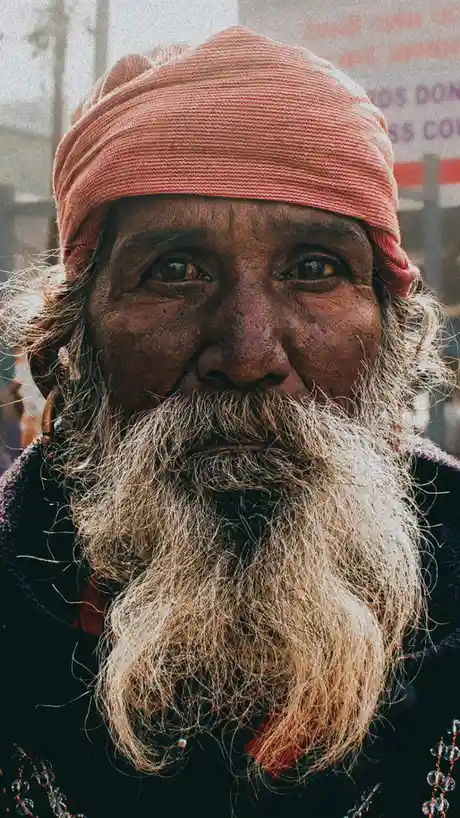 A close-up photo of an elderly man with a long, gray beard, wearing a pink headwrap and dark clothing, looking directly at…