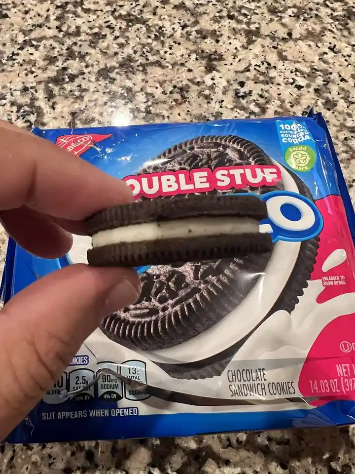 A hand holds a single chocolate sandwich cookie with white cream filling directly in front of its blue and pink packaging…