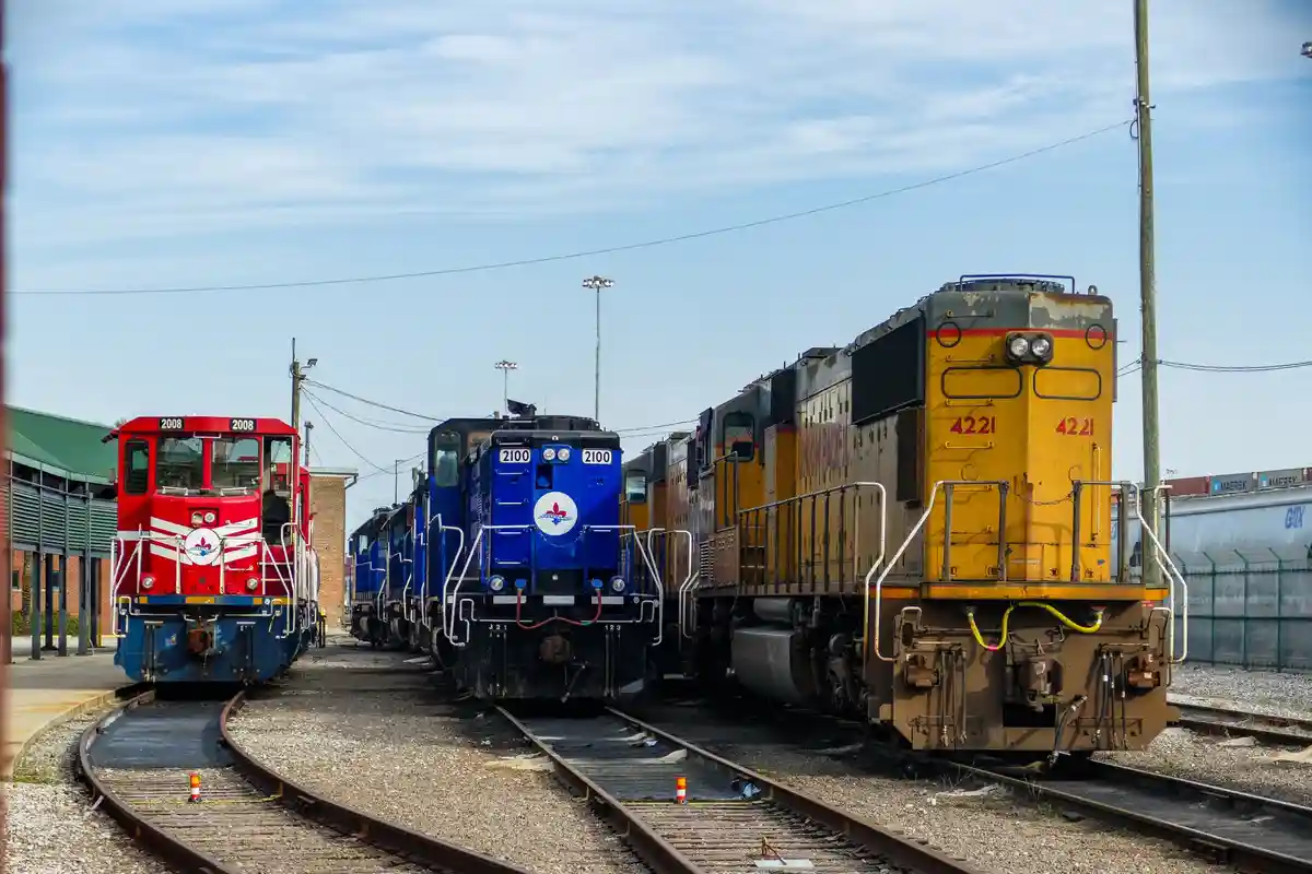 Three locomotives are parked side-by-side on separate tracks in a rail yard under a partly cloudy sky. The red engine on the…