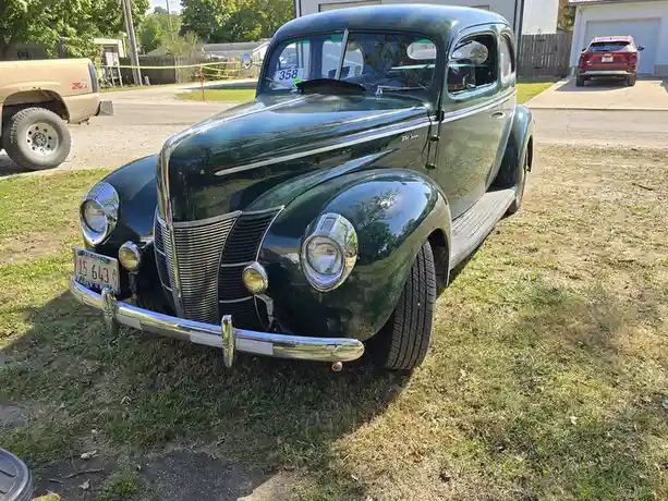 A shiny, dark green 1940 Ford Deluxe classic car is parked on grass, with a '358' auction tag on the windshield and an…
