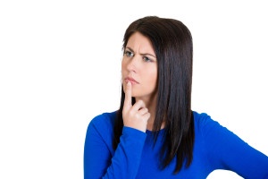 a woman with dark hair and a blue shirt looks confused and skeptical, her finger on her lips, against a white background…