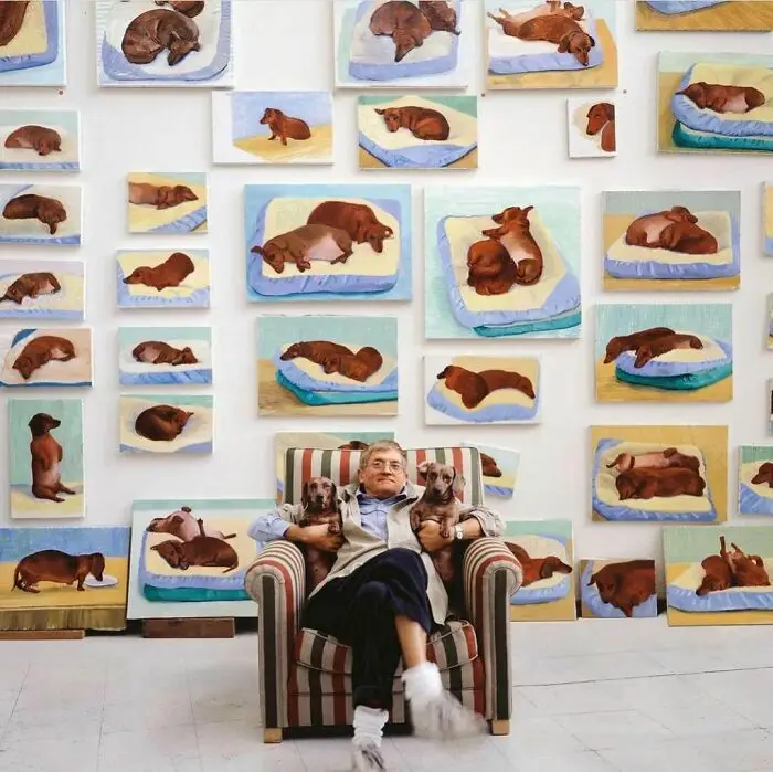A man sits in a striped armchair holding several dachshunds, surrounded by numerous paintings of dachshunds on beds. The…
