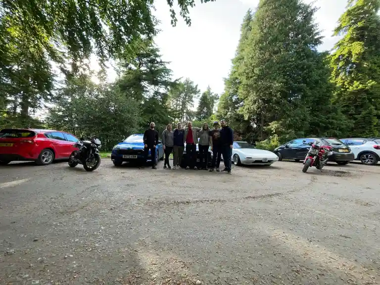 A group of seven people pose for a photo in a gravel parking lot surrounded by various cars and motorcycles, including a red…