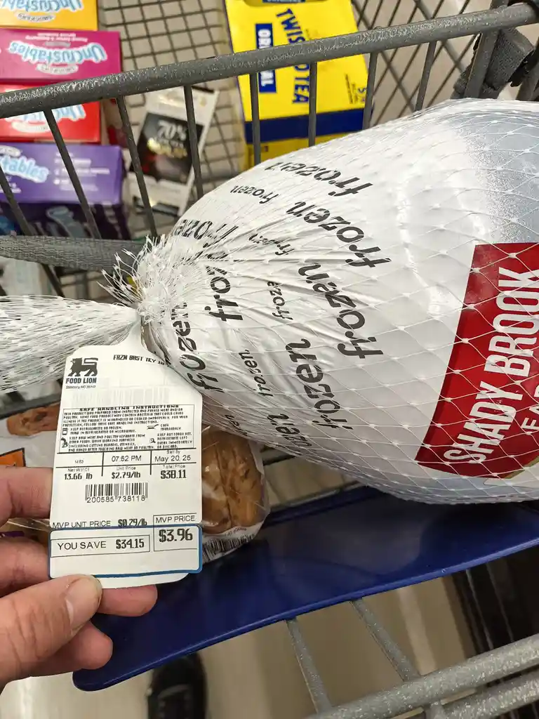 A hand holds a Food Lion price tag for a large frozen turkey in a shopping cart, showing a total price of $38.11 but a 'YOU…