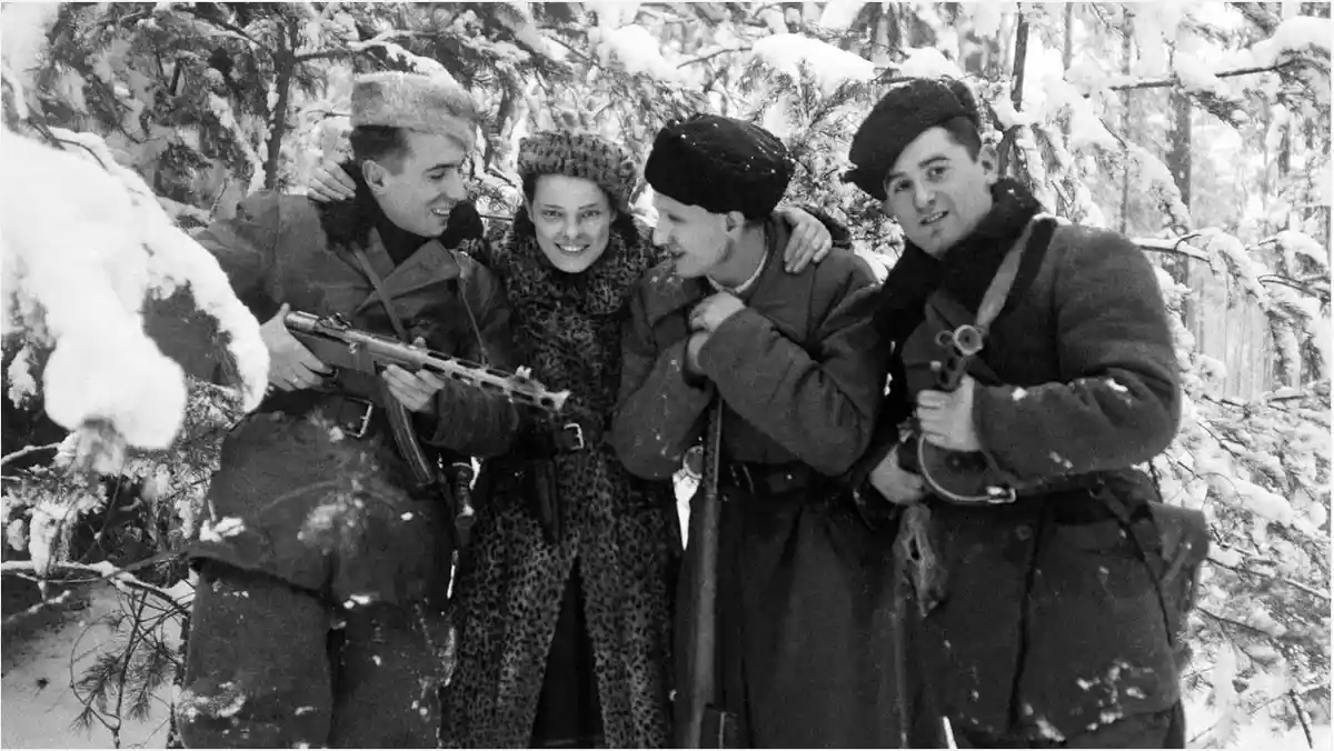 A black-and-white historical photo shows four people in winter military-style clothing standing in a snowy forest. Three…