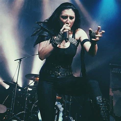 A female singer with long dark hair, wearing a black outfit with studded belt and arm accessories, is performing on stage…
