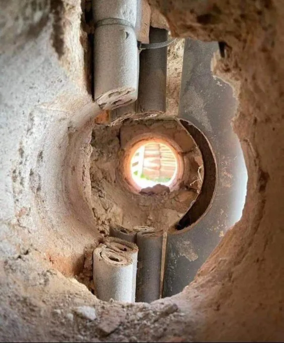 A close-up view from inside a rough, circular hole in a concrete or earthen wall, looking through a broken pipe opening…