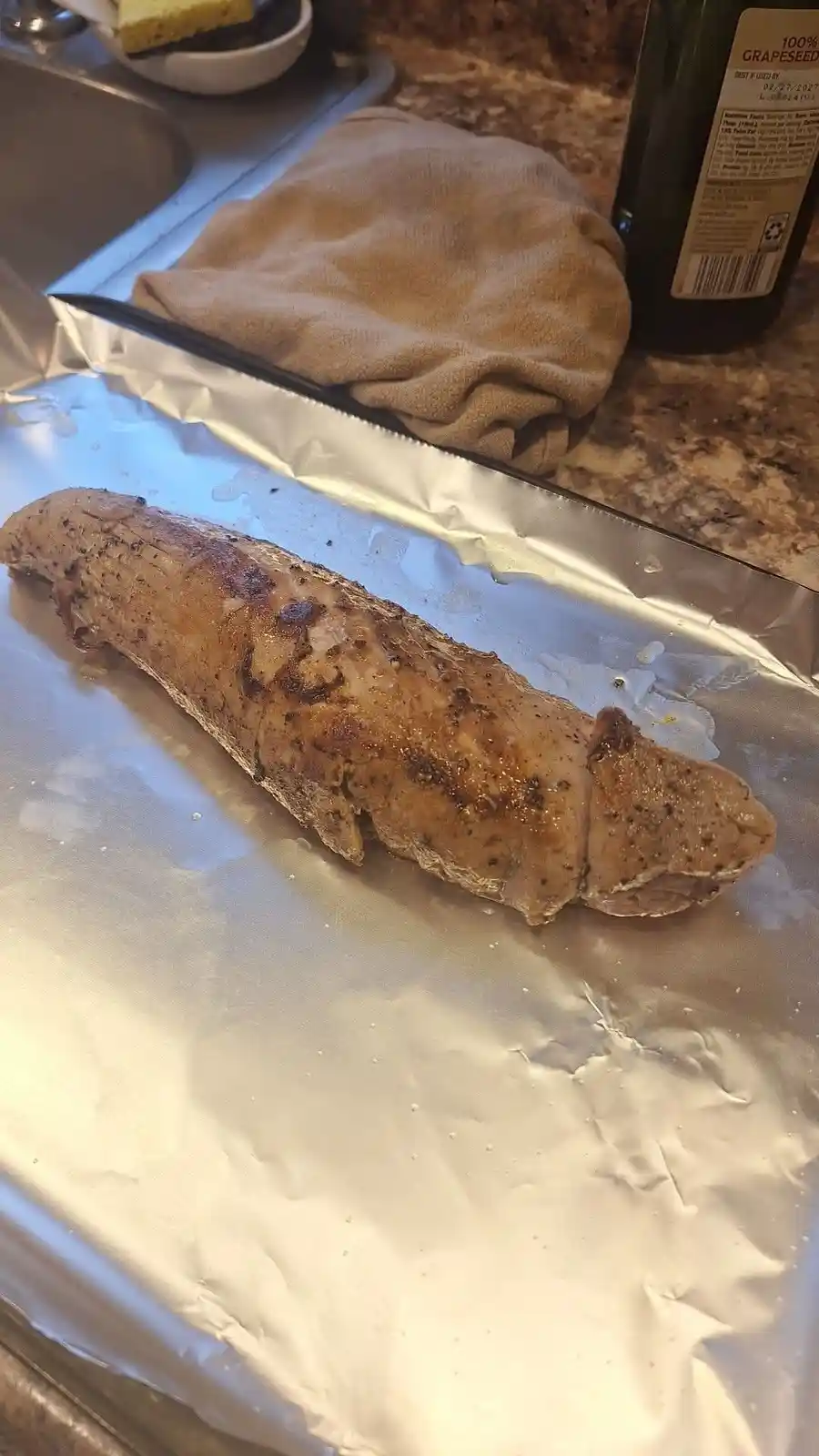 A cooked, long, cylindrical piece of meat, resembling a sausage, rests on aluminum foil on a baking tray. A large knife lies…