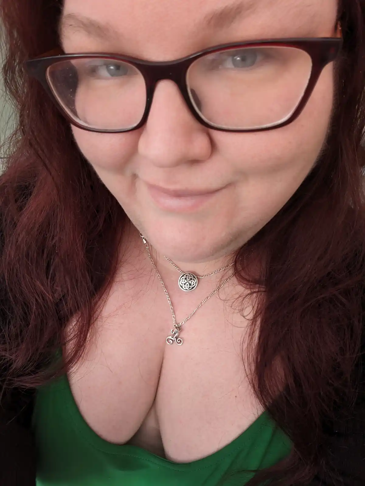 A close-up photo of a woman with reddish-brown hair and glasses, wearing a green top and two silver necklaces. One necklace…