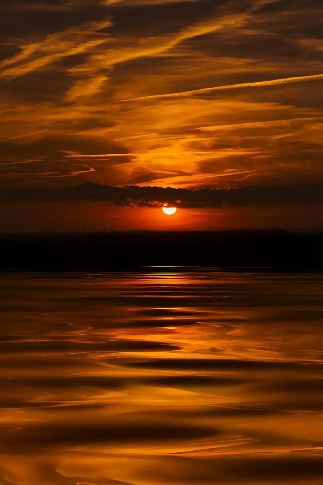 A serene sunset over water, with the sun partially obscured by clouds, casting vibrant orange and yellow hues across the sky…