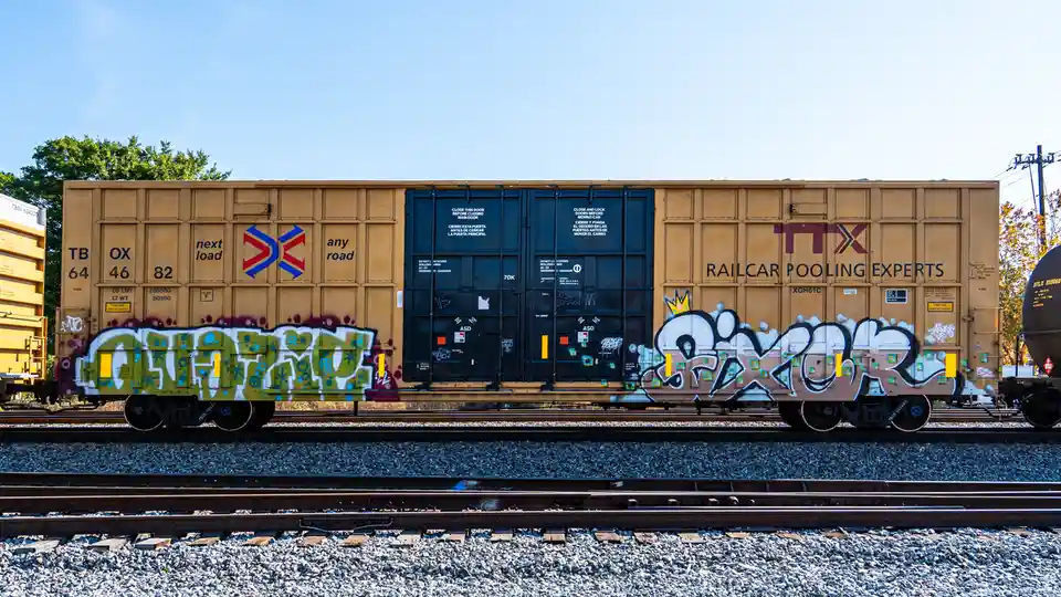 A yellow freight train car with 'TTX RAILCAR POOLING EXPERTS' branding and graffiti art, including a large white and brown…