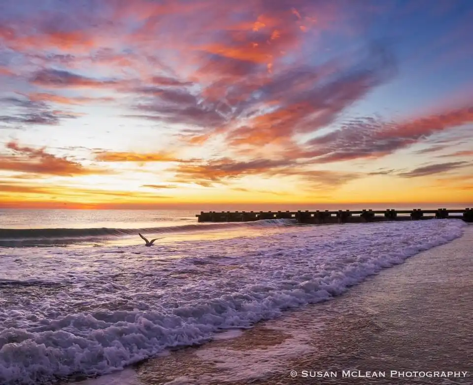 A serene beach sunset photo with vibrant orange, pink, and purple clouds over the ocean, waves crashing on shore, and a…