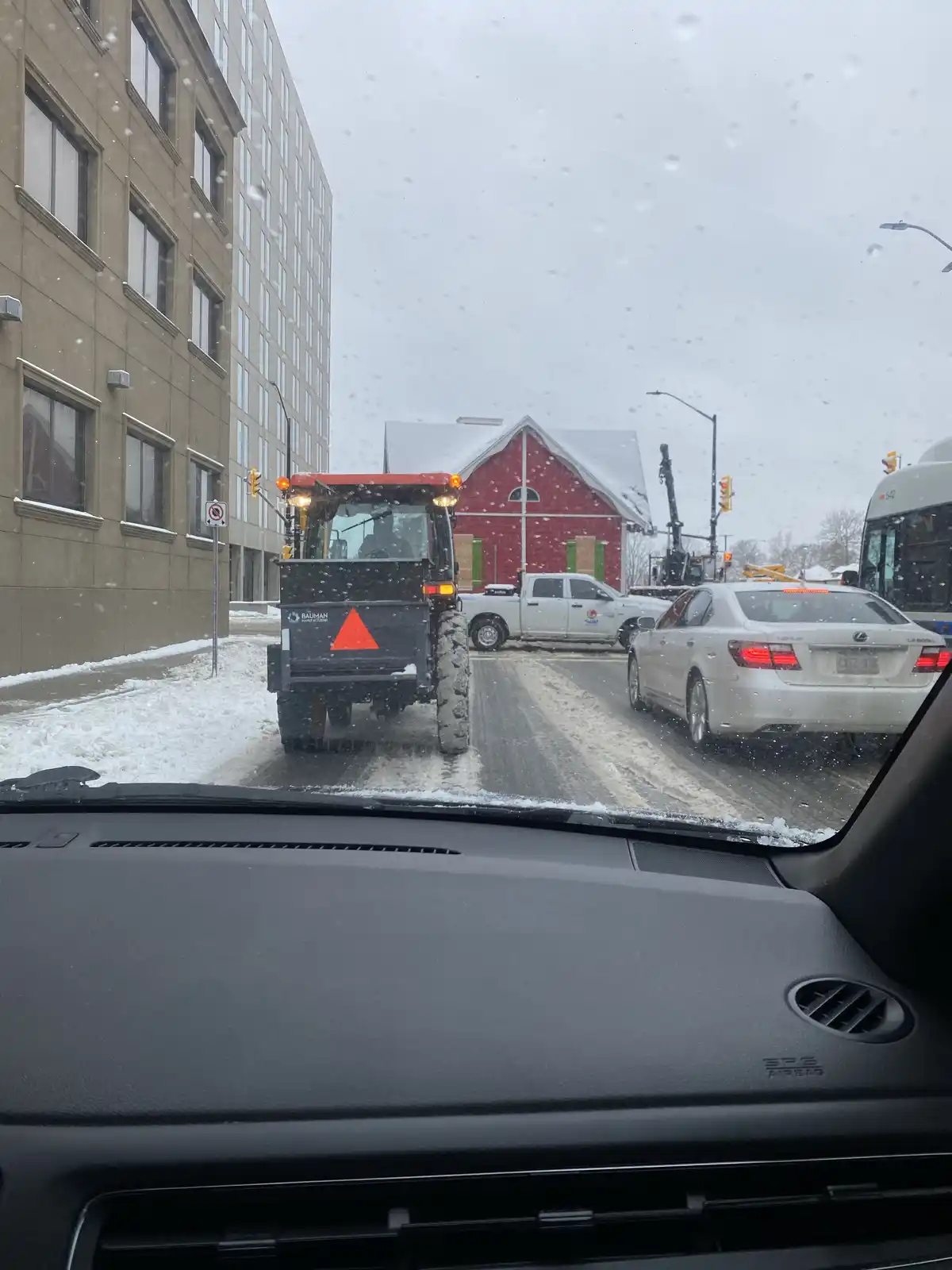 A snowy urban street scene viewed from inside a car, showing a red and black snowplow tractor stopped in traffic alongside a…