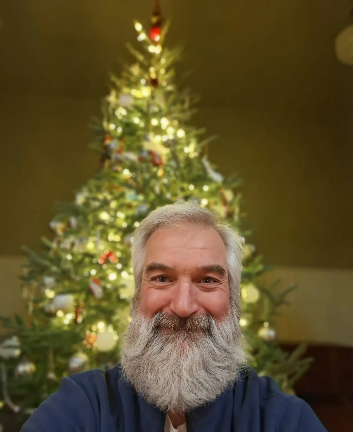 A man with a long white beard and gray hair smiles at the camera, wearing a blue hoodie. Behind him, a decorated Christmas…