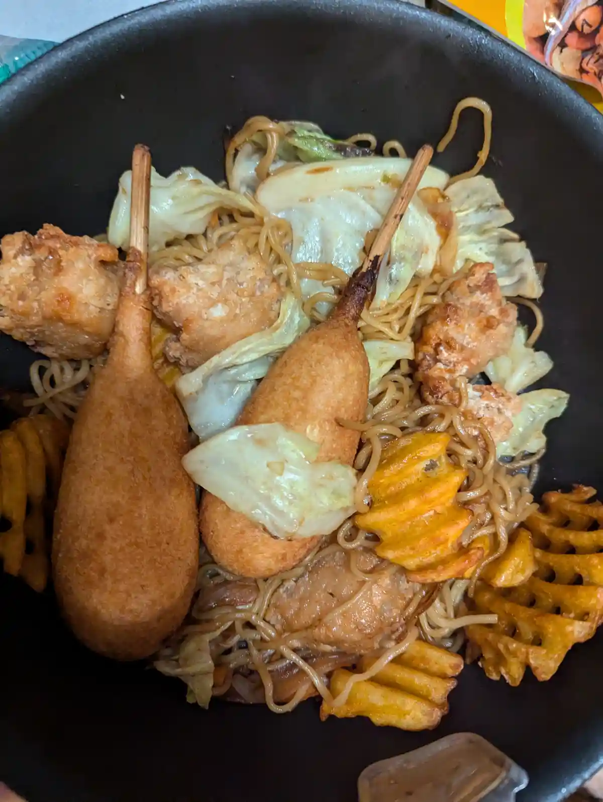 A black bowl contains a chaotic mix of ramen noodles, fried chicken pieces, waffle fries, and two corn dogs on sticks, all…