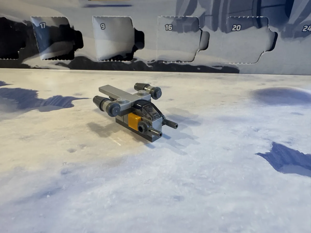 A small, gray and yellow Lego X-wing fighter model sits on a snowy surface in front of a partially opened Lego Advent…