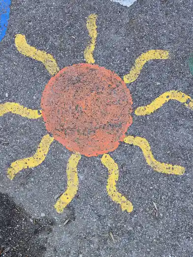A childlike drawing of a sun painted on asphalt, featuring an orange circle with uneven yellow rays. The overlaid text reads…