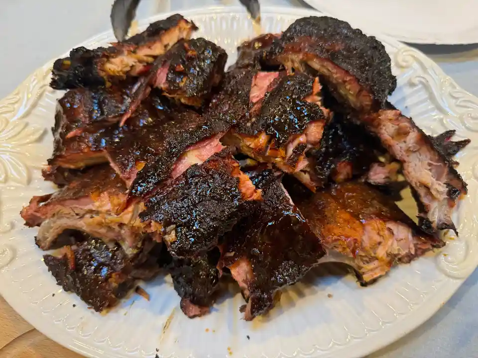 A white ornate plate is piled high with heavily charred, dark-brown smoked ribs, some pieces showing pinkish meat and bone…
