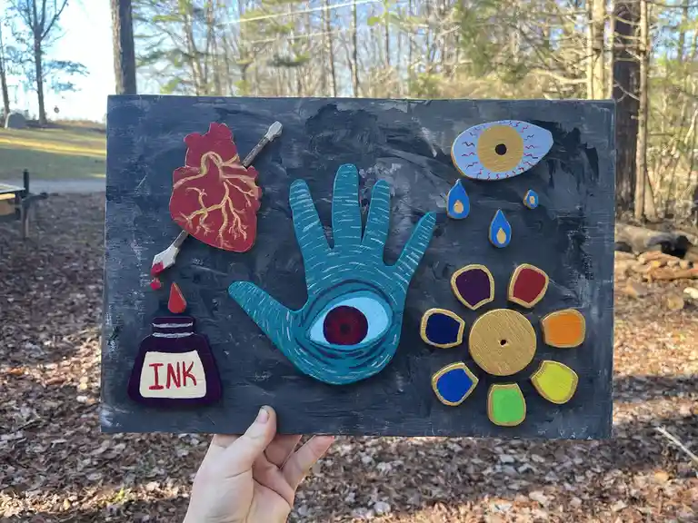 A hand holds a dark wooden plaque featuring various painted wooden cutouts: a red heart pierced by a paintbrush, a blue hand…