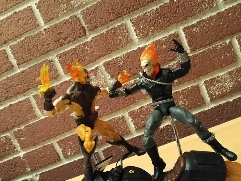 Two Ghost Rider action figures are posed in a dynamic fighting stance against a brick wall background, one with orange flame…