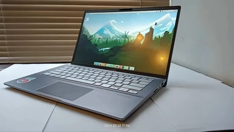 A silver laptop sits open on a white desk with a mountainous desktop wallpaper, featuring a sticker on the palm rest and a…