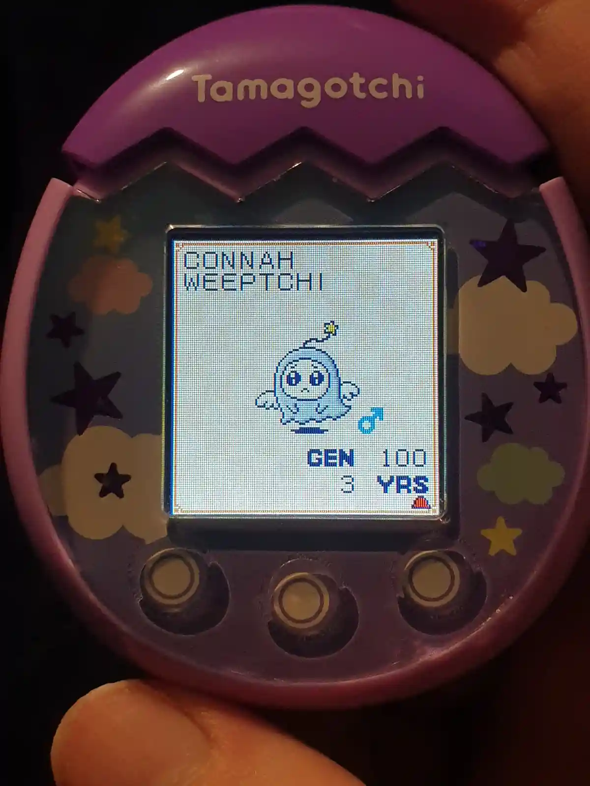 A close-up of a purple Tamagotchi handheld device showing a pixelated blue Weeptchi digital pet with a sad expression. The…