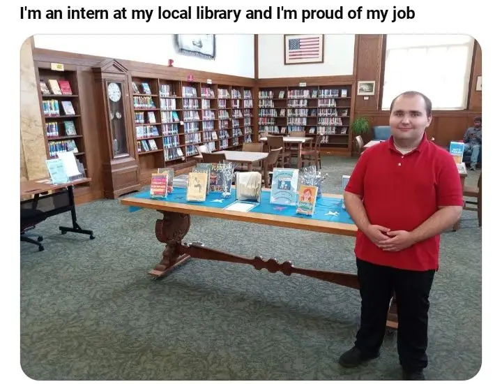 A man in a red polo shirt stands confidently in a library, smiling at the camera with his hands clasped. Behind him are…