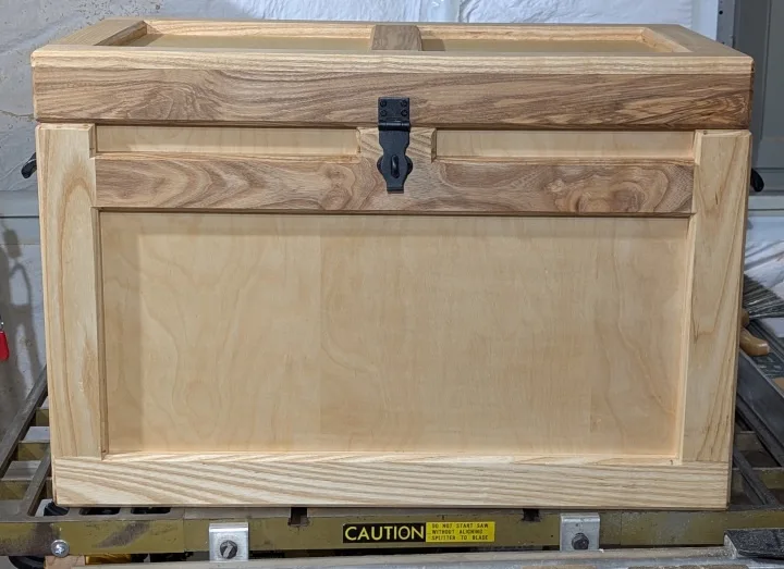 A wooden chest with a black latch sits on a workbench, and a yellow caution sticker on the bench reads 'CAUTION DO NOT START…